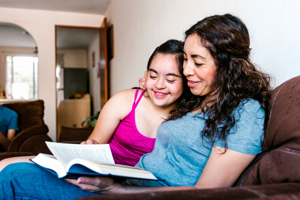 Smiling latin teen girl with down syndrome and her mom reading a book at home, in disability concept in Latin America